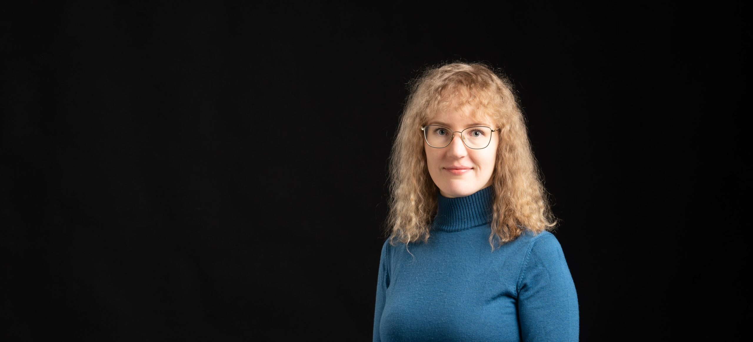 Kuvassa oikeassa reunassa on vaalea kiharatukkainen henkilö, jolla on yllään sininen paita. Kuvan tausta on täysin musta.