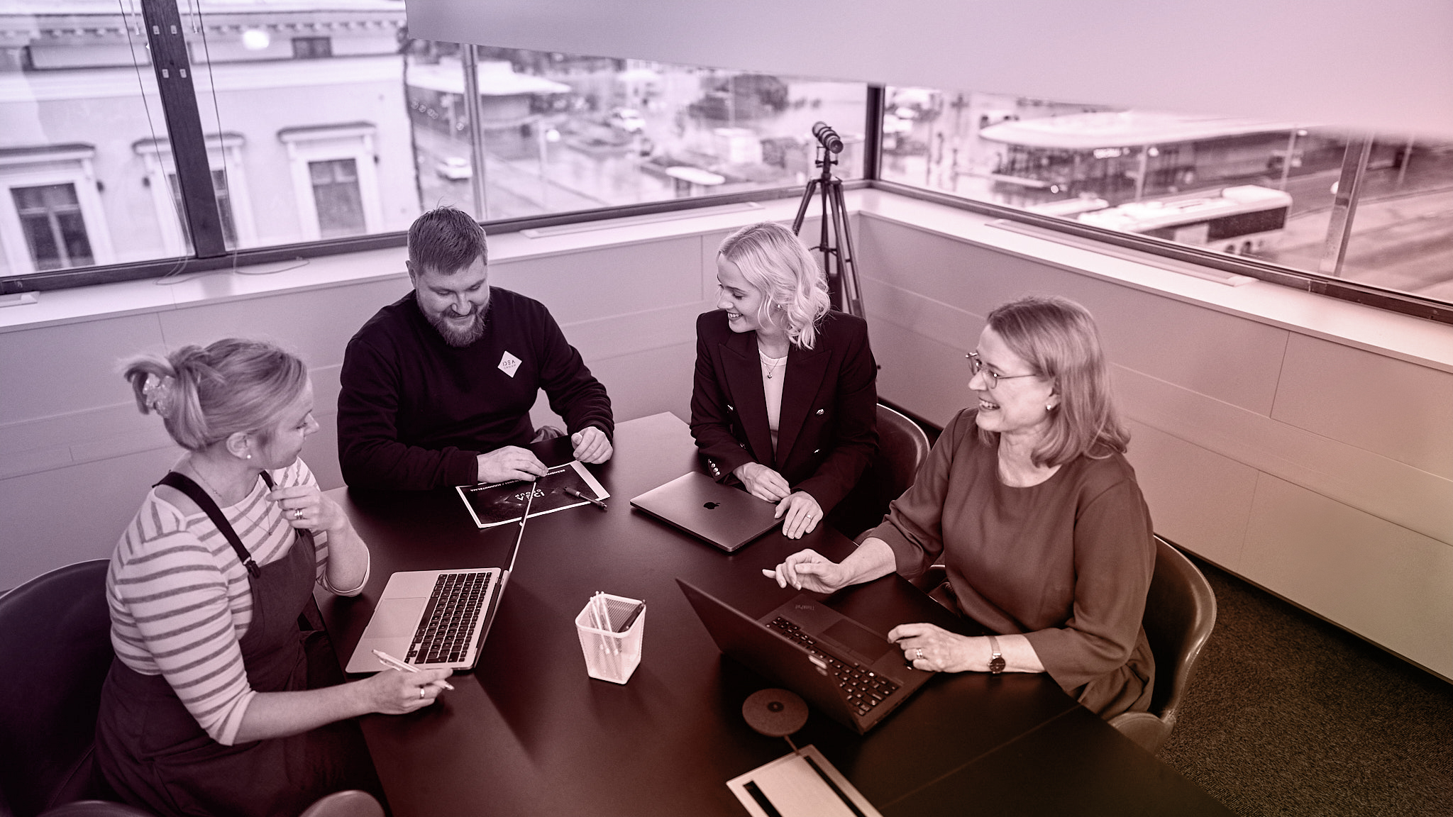 Mies ja kolme naista istuvat neuvottelupöydän ääressä. Pöydällä on tietokoneita, taustalla ikkunasta näkyy tori.
