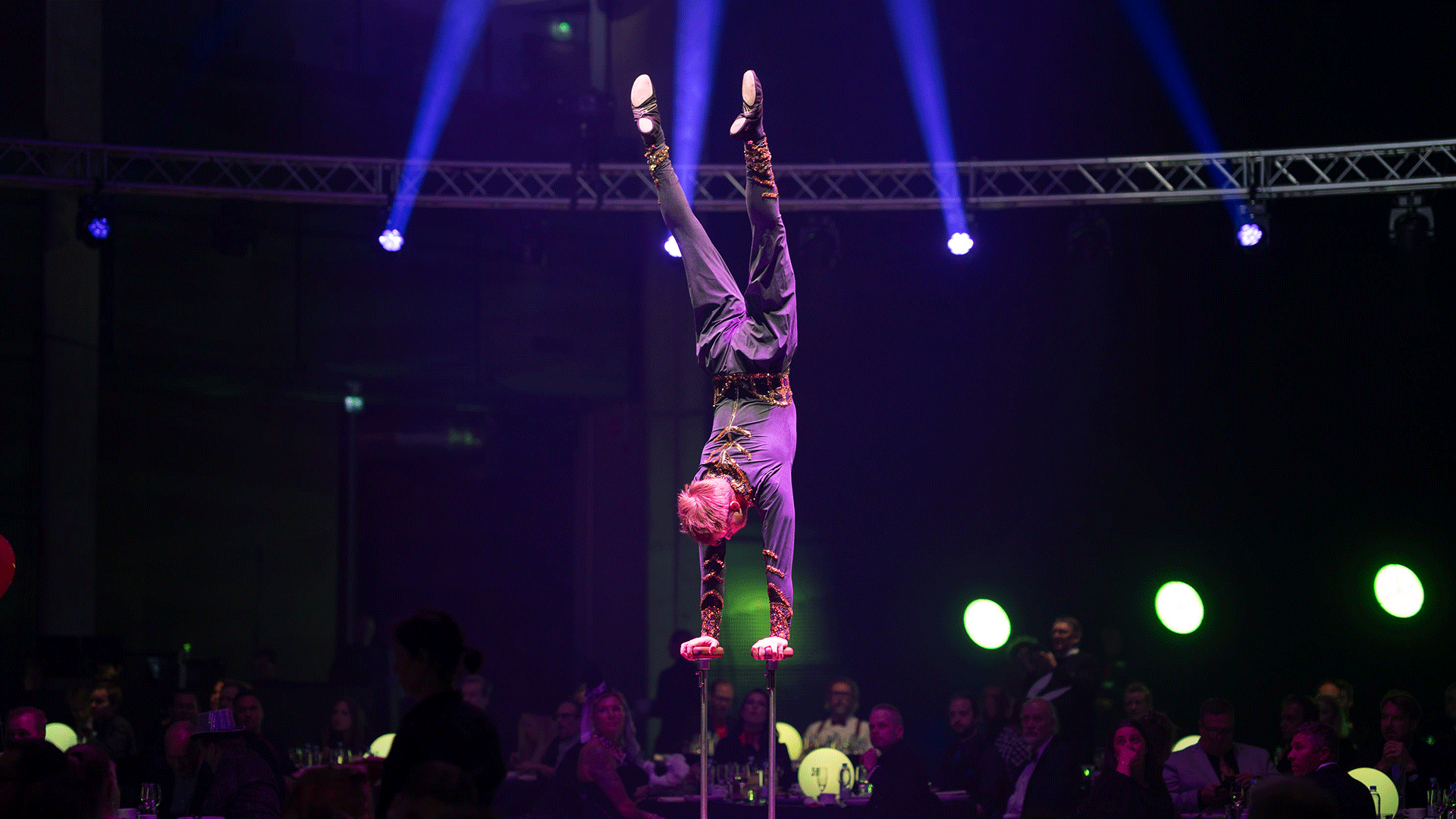 Sirkustaiteilja seisoo käsillään seisontatukien päällä, taustalla on sinisiä spottivaloja