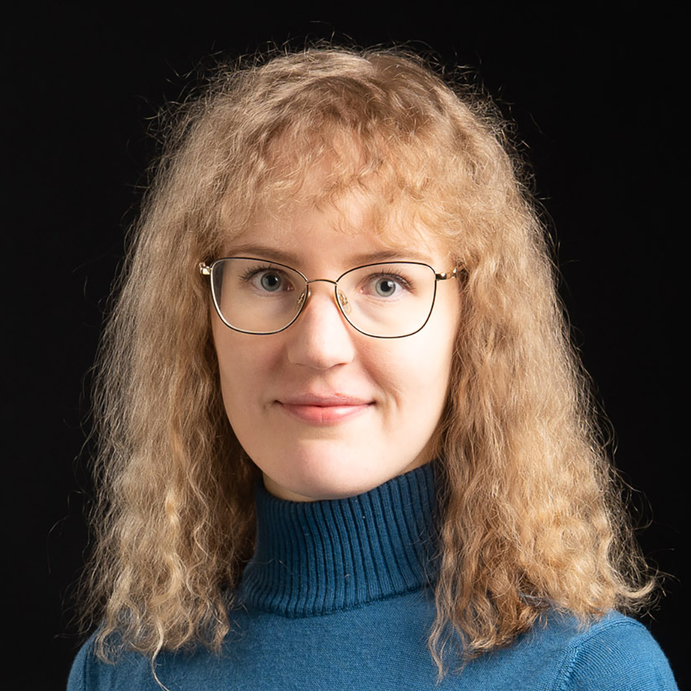 Kuvassa keskellä on vaalea kiharatukkainen henkilö, jolla on yllään sininen paita. Kuvan tausta on täysin musta.